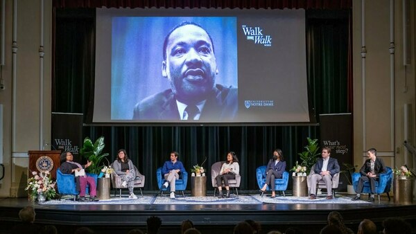 Seven people sitting on chairs on a stage below a projection of MLK Jr.