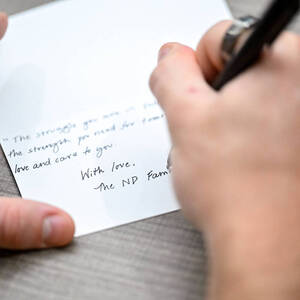 A hand holding a pen writes an encouraging note on a notecard