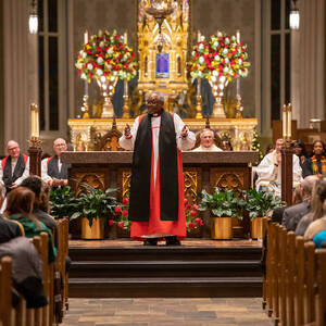 Bishop Michael Curry stands at the front of the Basilica, in the center aisle, with his arms spread wide, and speaks to the audience