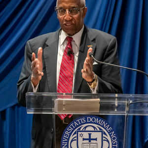 Howard Adams, an African American man in a suit and tie, stands at a podium and gestures with his hands