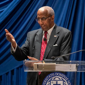 Howard Adams, an African American man in a suit and tie, stands at a podium and gestures with his right hand