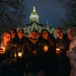 A group of students and staff wear winter coats outside on snowy evening while holding candles. The main building is behind them.
