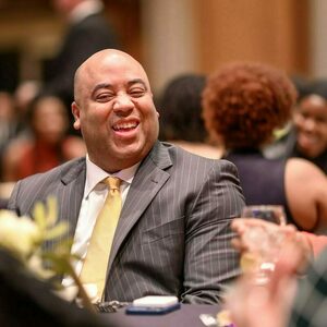 A bald black man in a suit and tie smiles as he sits at a dinner table