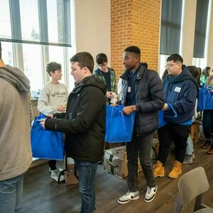 a line of students holding blue fabric bags walk past a table
